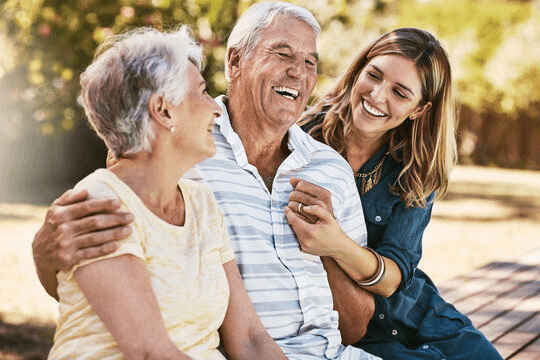 A grown-up daughter laughing with her elderly mother and father, her arm around her father, outdoors on a sunny day