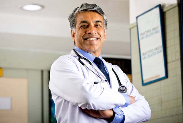 A smiling doctor in a white coat, stethoscope around his neck, standing with his arms folded in his clinic